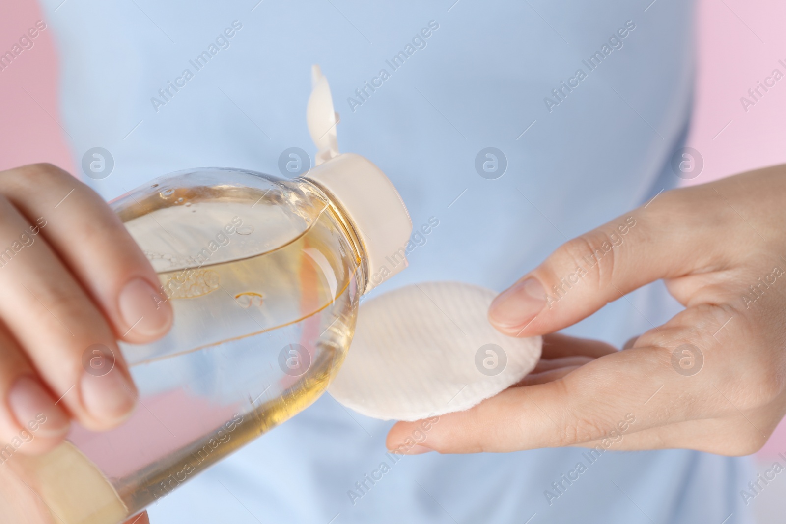 Photo of Woman pouring micellar water from bottle on cotton pad against pink background, closeup