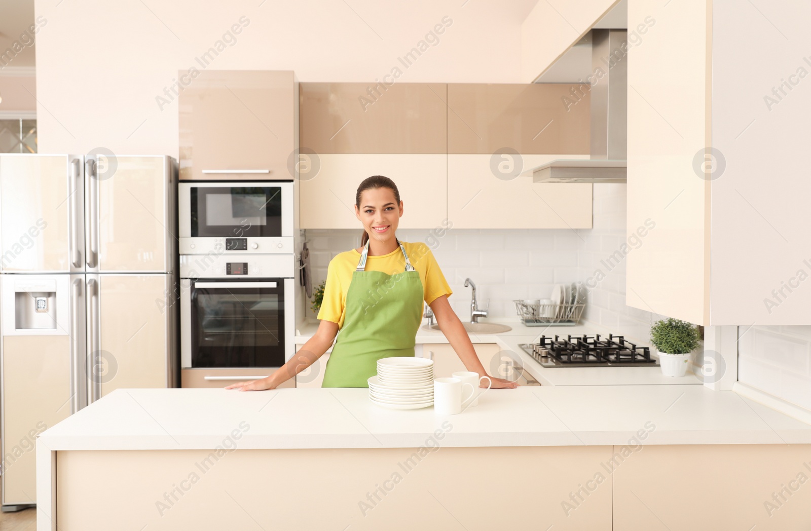 Photo of Beautiful young woman with clean dishes and cups at table in kitchen