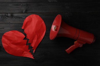 Photo of Torn red paper heart and megaphone on black wooden background, flat lay. Relationship problems concept
