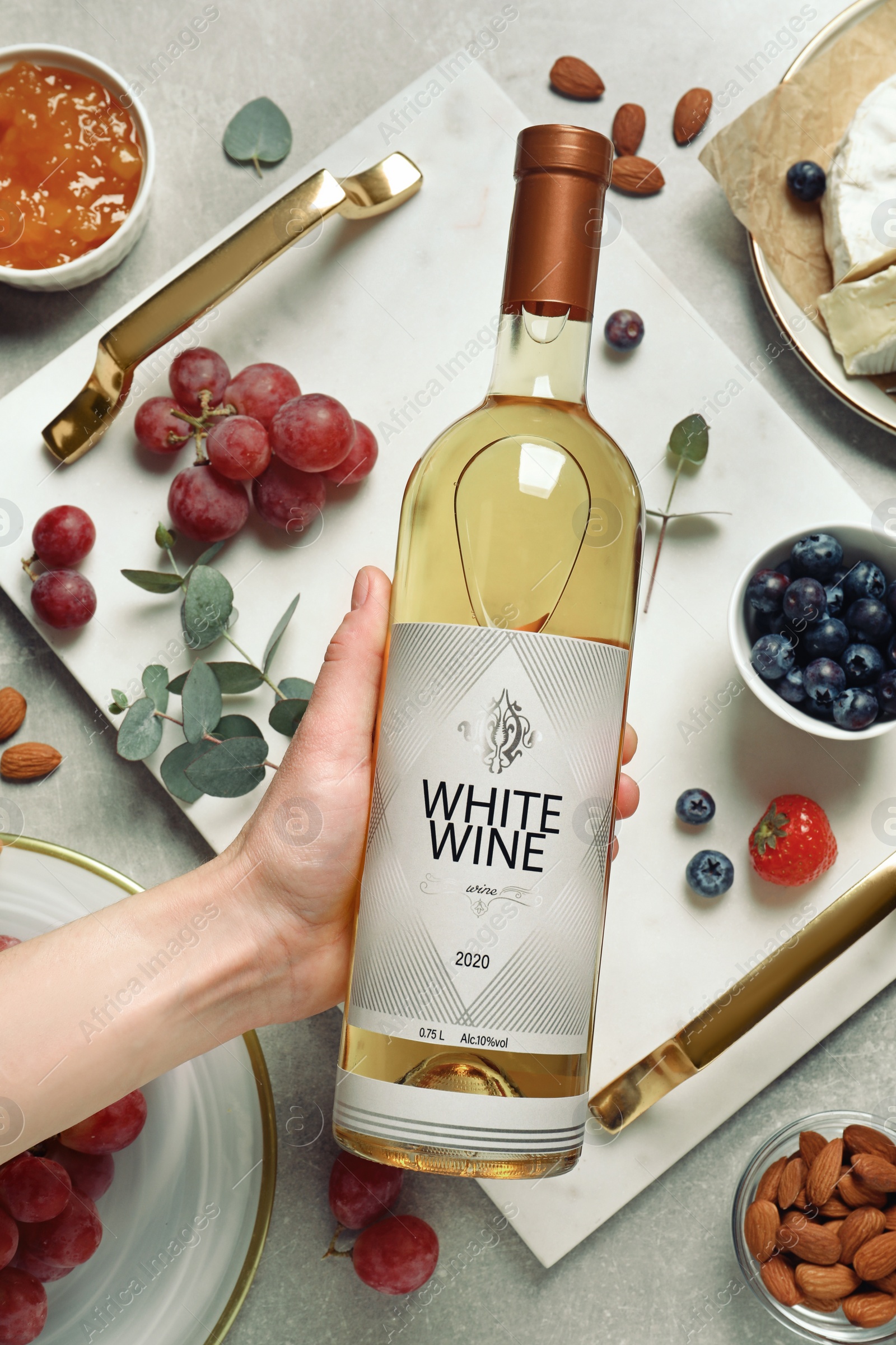 Photo of Woman holding bottle of white wine at grey table with snacks, top view