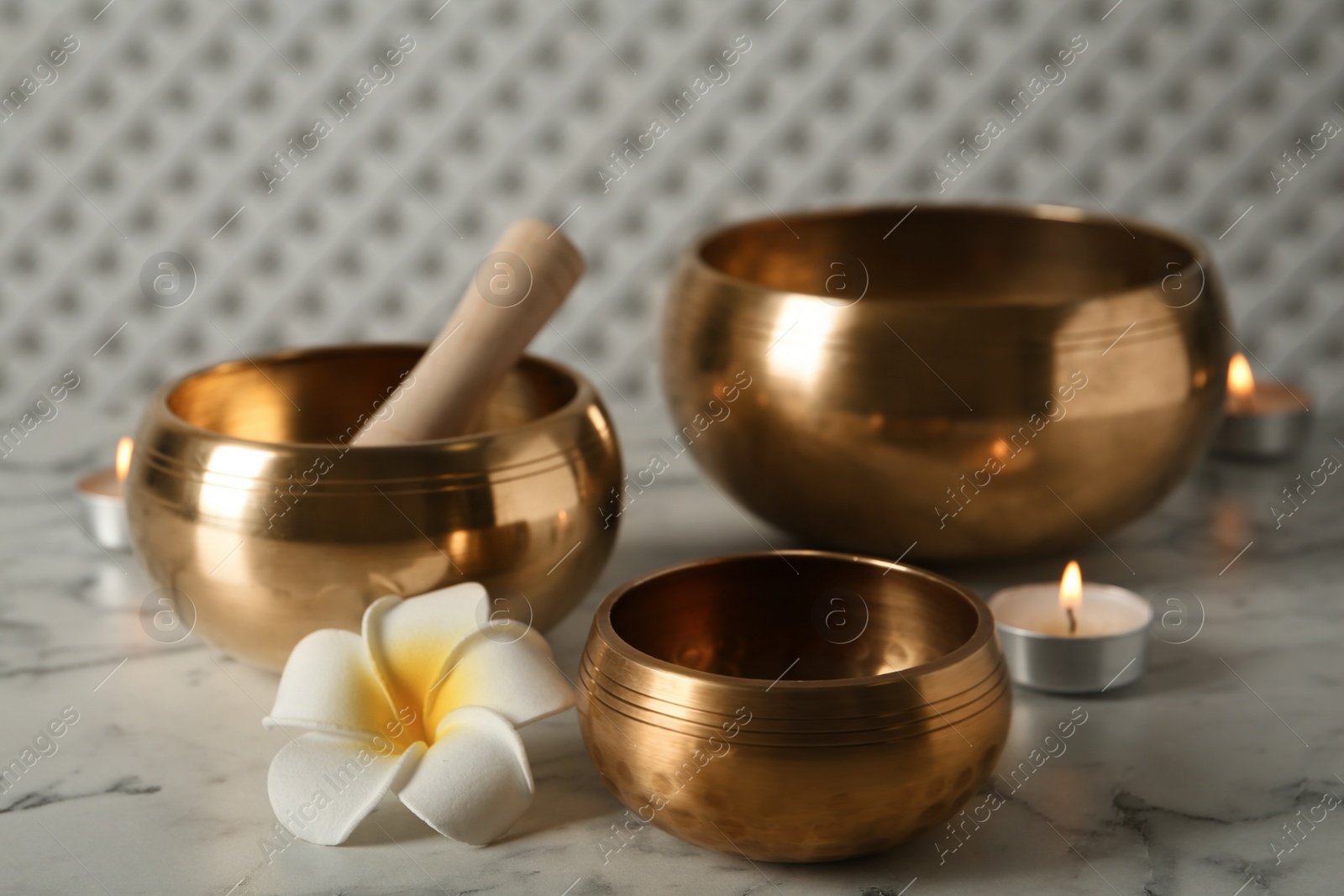 Photo of Tibetan singing bowls, mallet, plumeria flower and burning candles on white marble table