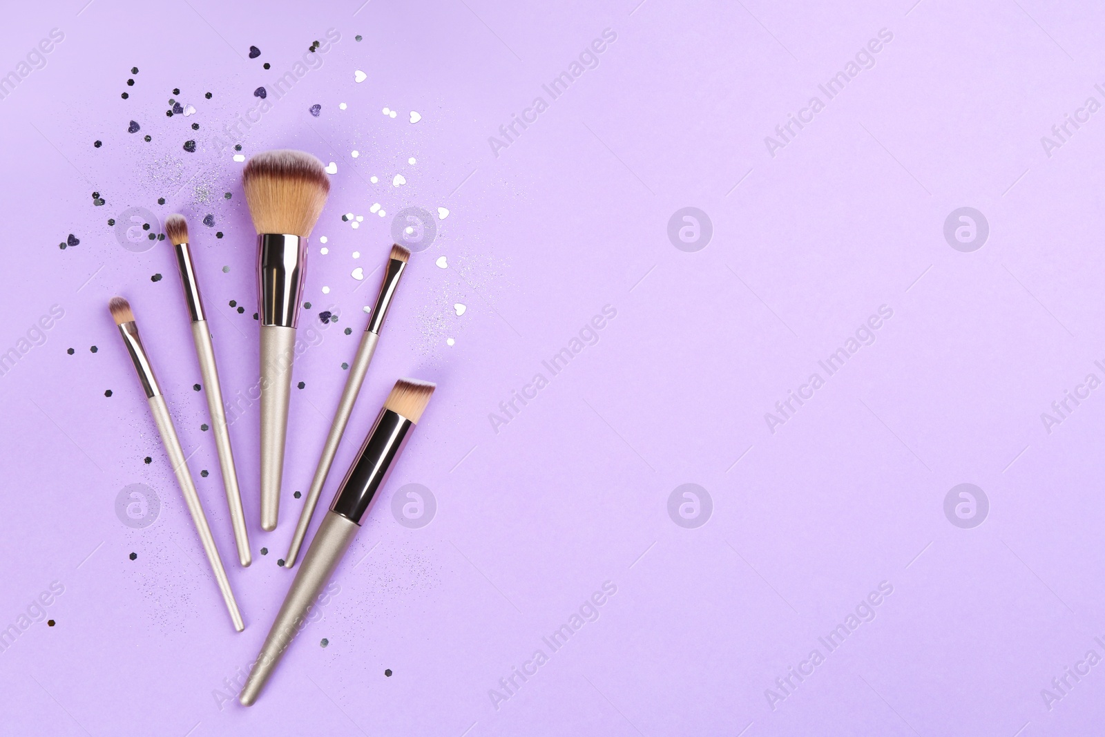 Photo of Different makeup brushes and shiny confetti on violet background, flat lay. Space for text