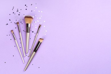 Different makeup brushes and shiny confetti on violet background, flat lay. Space for text