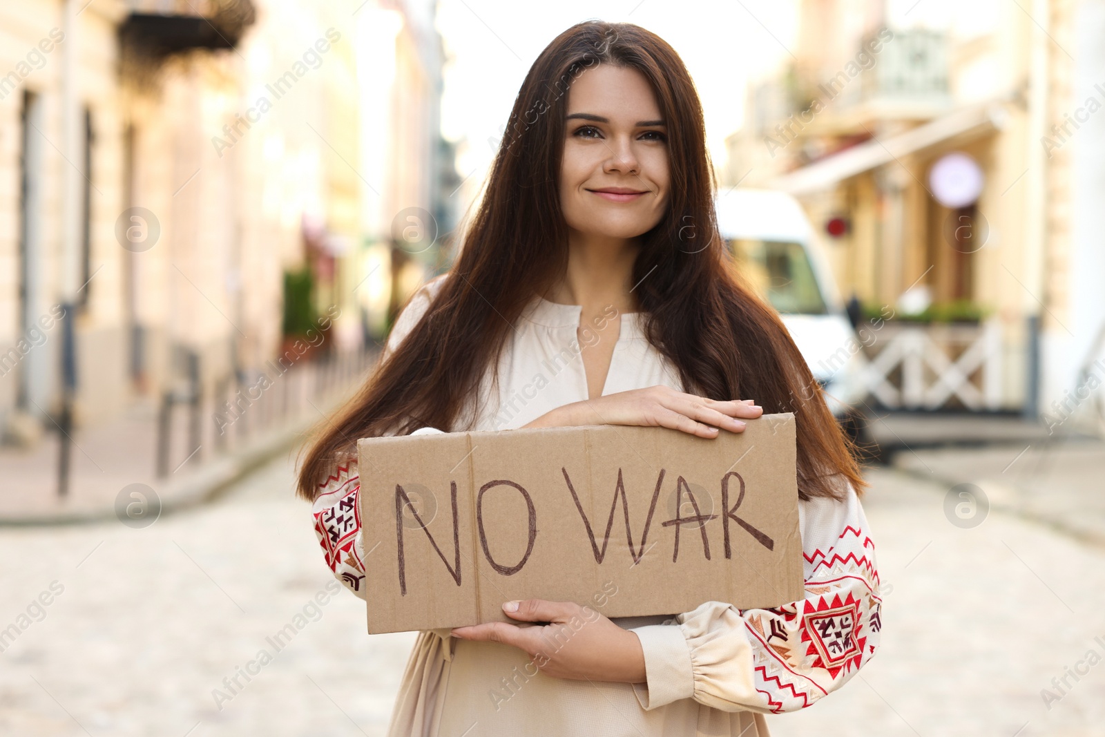 Photo of Smiling woman in embroidered dress holding poster No War on city street