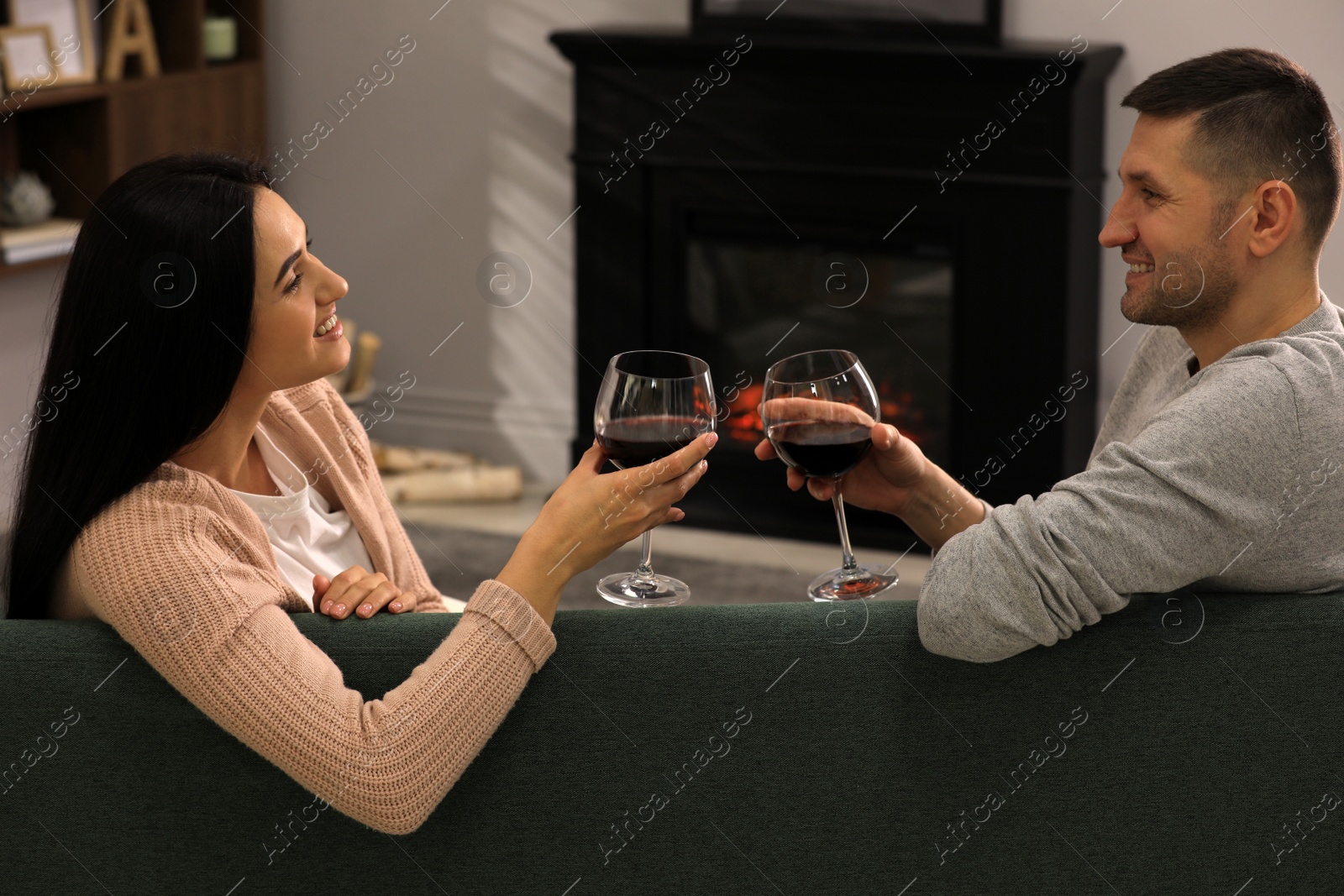 Photo of Lovely couple with glasses of wine spending time together on sofa near fireplace at home