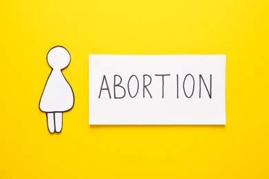 Photo of Note with word Abortion and pregnant woman paper cutout on yellow background, flat lay