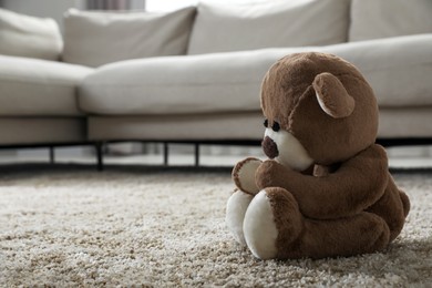 Cute lonely teddy bear on floor near sofa in room. Space for text