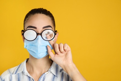 Woman wiping foggy glasses caused by wearing medical mask on yellow background. Space for text