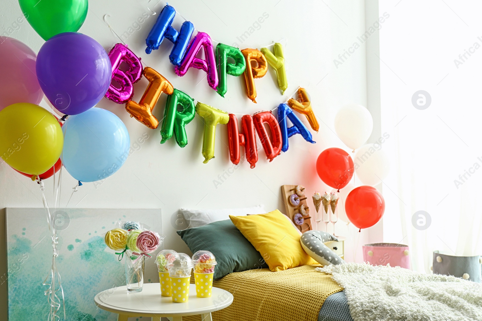 Photo of Phrase HAPPY BIRTHDAY made of colorful balloon letters and table with treats in modern bedroom