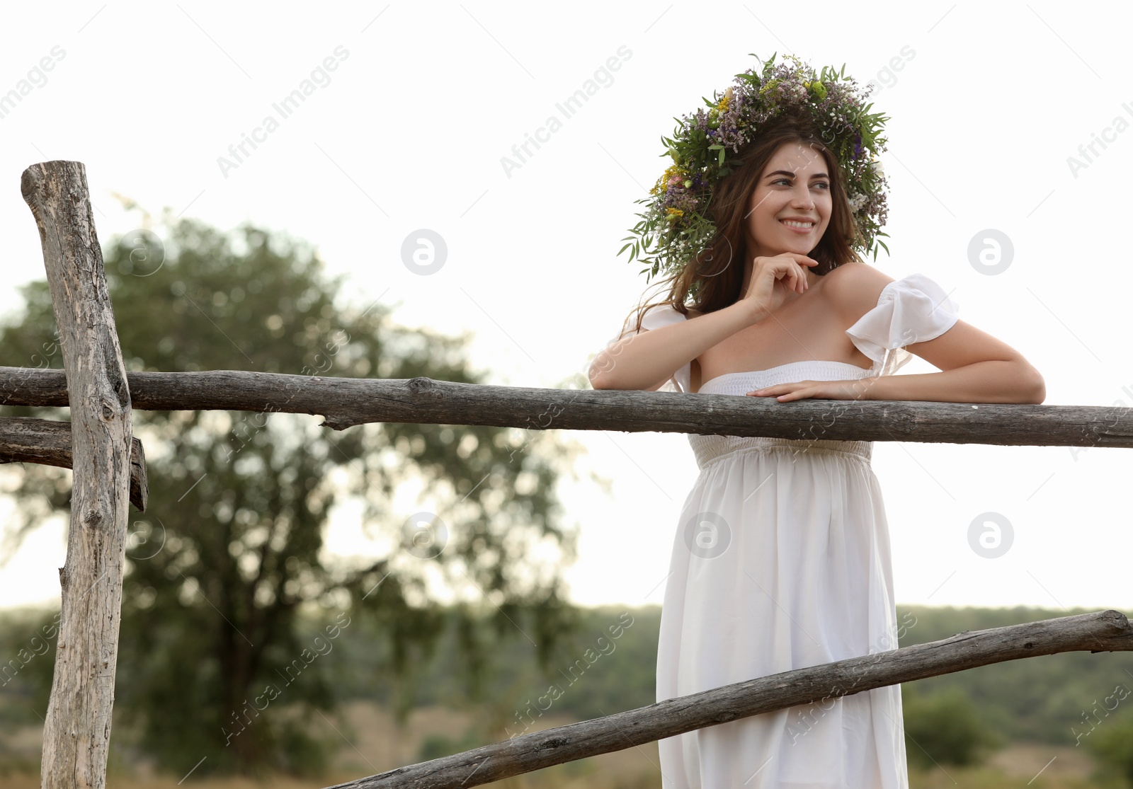 Photo of Young woman wearing wreath made of beautiful flowers near wooden fence