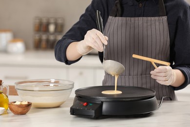 Photo of Woman cooking delicious crepe on electric pancake maker at white marble table in kitchen, closeup