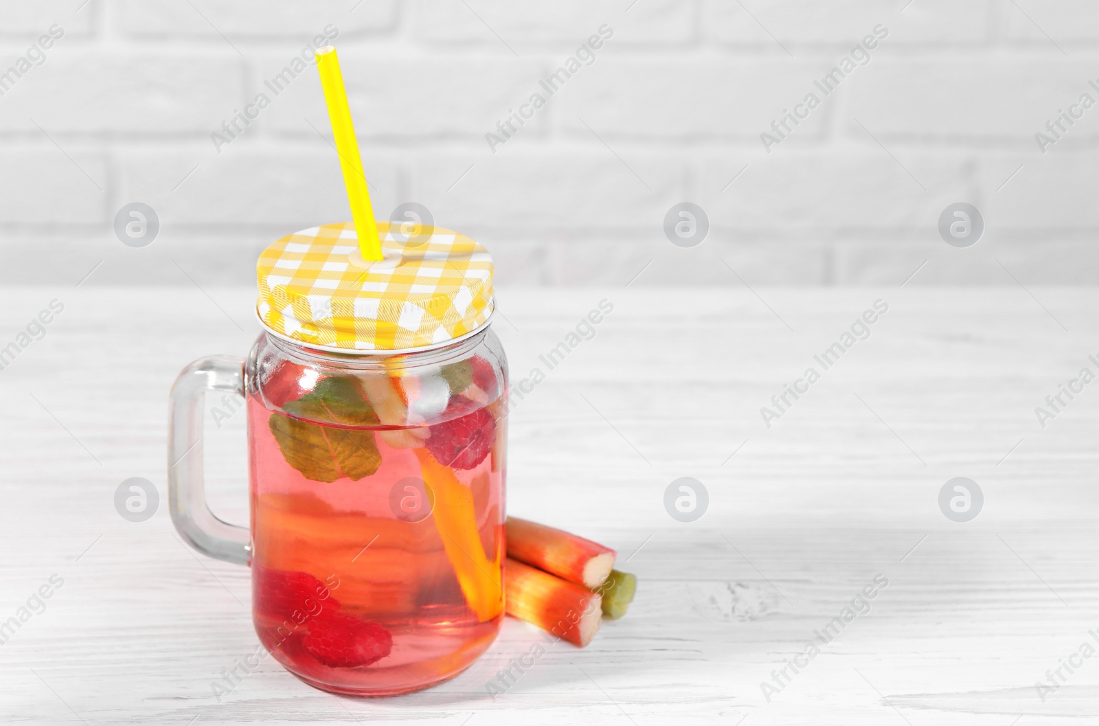 Photo of Mason jar of tasty rhubarb cocktail with raspberry and stalks on white wooden table. Space for text