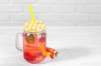 Mason jar of tasty rhubarb cocktail with raspberry and stalks on white wooden table. Space for text