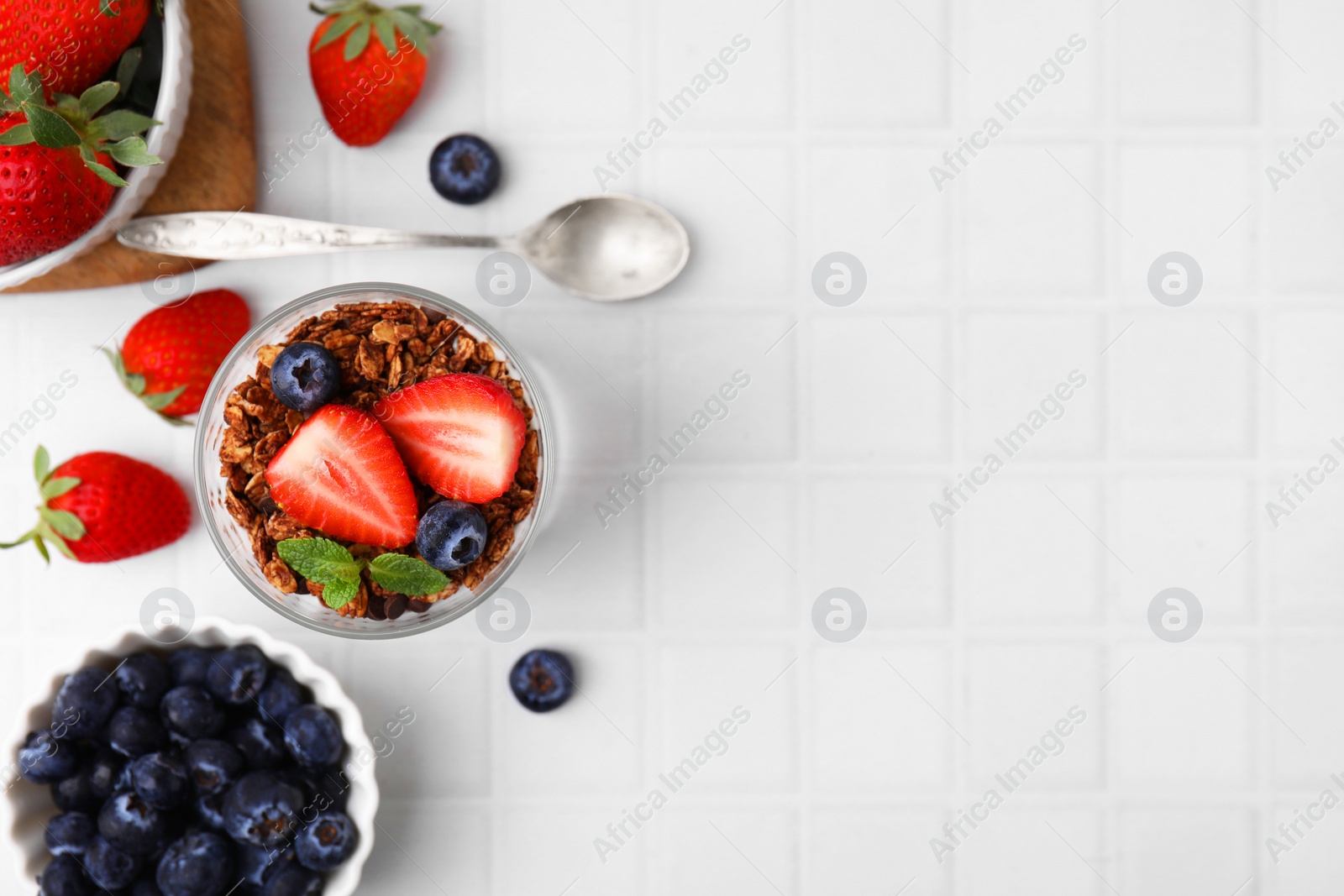 Photo of Tasty granola, strawberries, blueberries and spoon on white tiled table, flat lay. Space for text