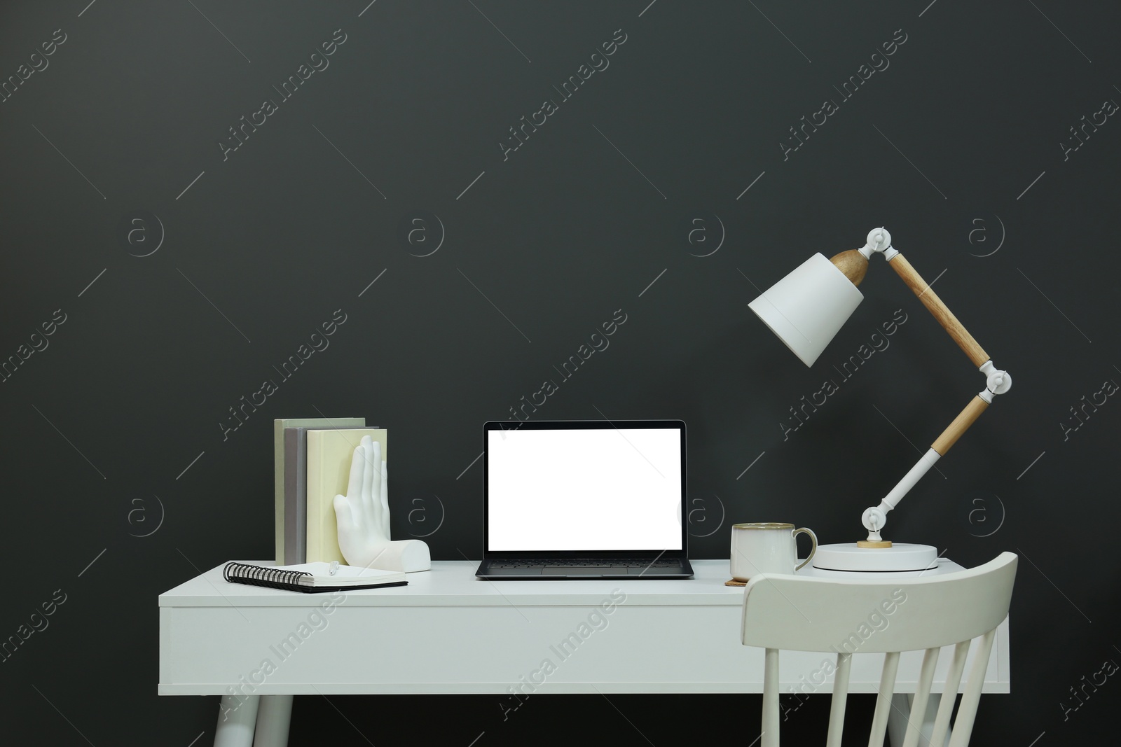 Photo of Stylish workplace with laptop, lamp, cup and decor on white table near grey wall
