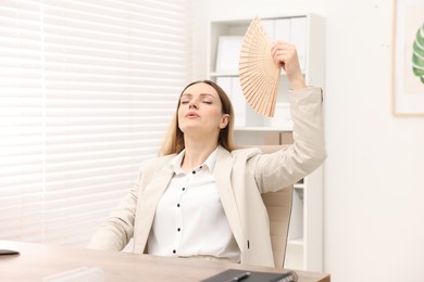 Photo of Businesswoman waving hand fan to cool herself at table in office