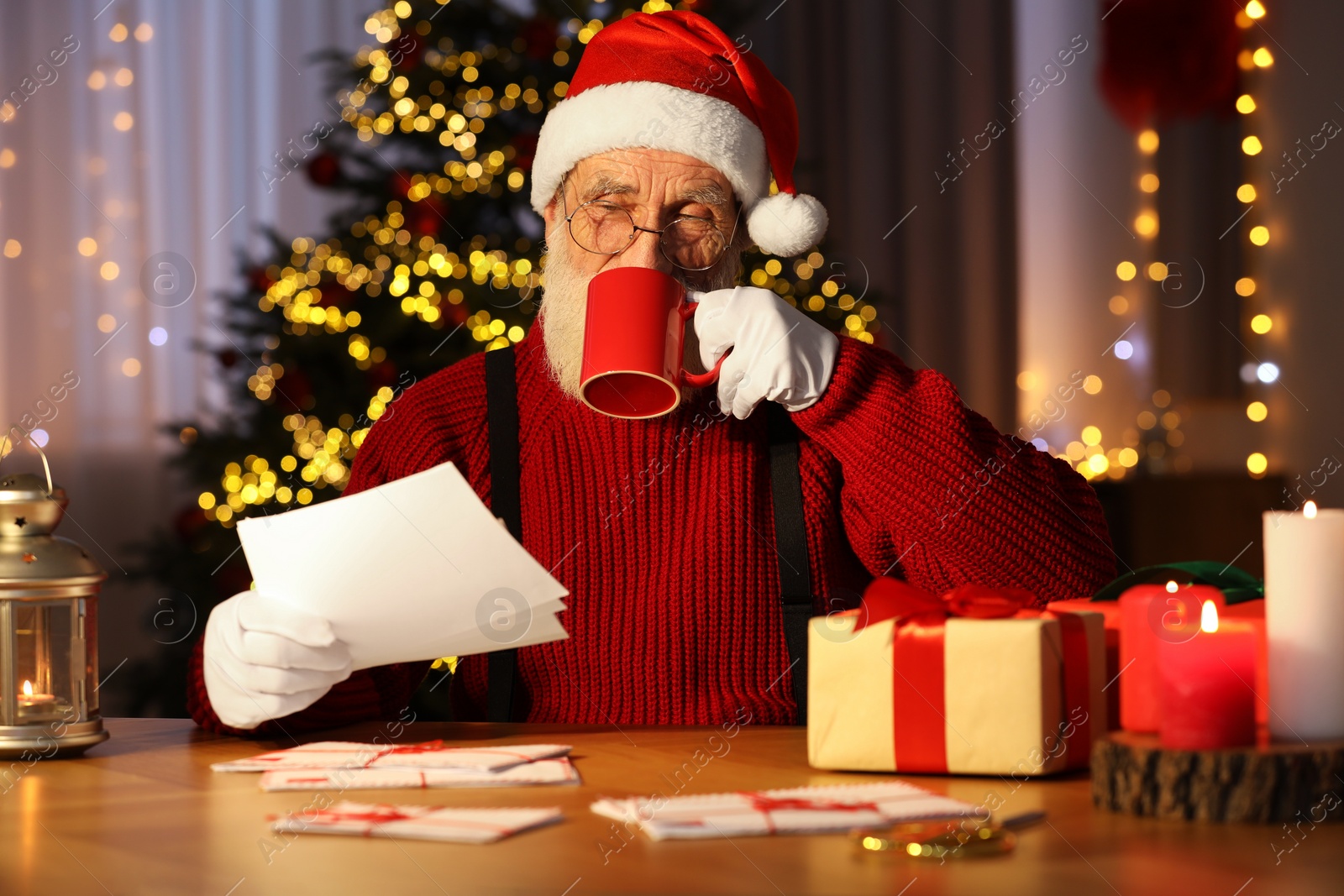 Photo of Santa Claus reading letter and drinking hot beverage at his workplace in room decorated for Christmas