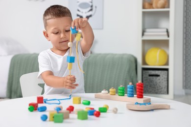 Motor skills development. Little boy playing with wooden pieces and string for threading activity at white table indoors