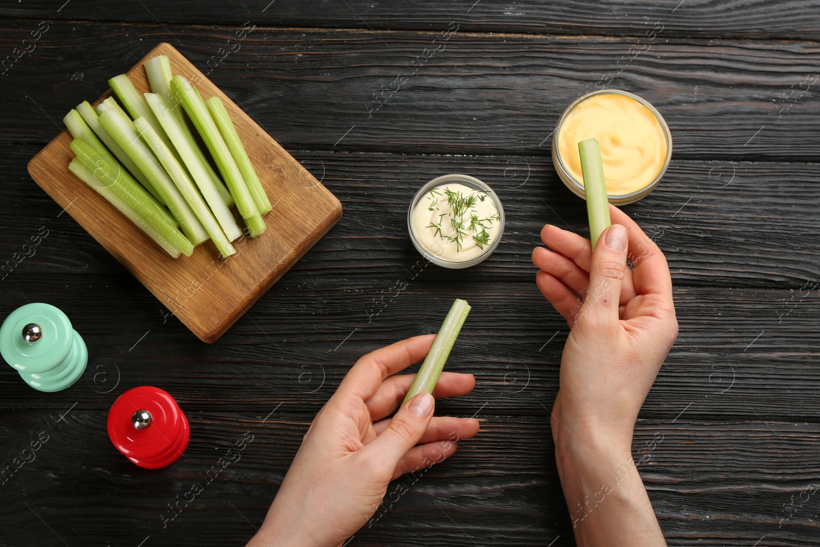 Photo of Woman dipping celery stick in sauce at dark wooden table, top view