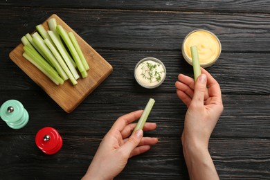 Photo of Woman dipping celery stick in sauce at dark wooden table, top view
