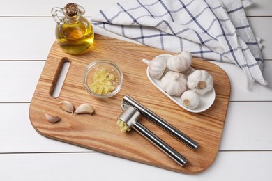 Photo of Board with metal press, oil and garlic on white wooden table, top view