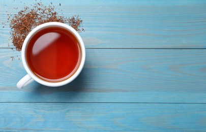 Photo of Ceramic cup of aromatic rooibos tea and scattered dry leaves on light blue wooden table, flat lay. Space for text