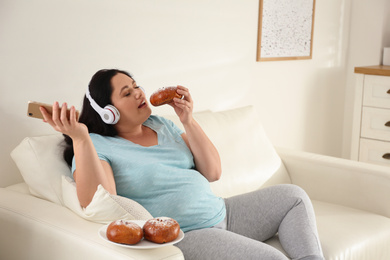 Photo of Lazy overweight woman with mobile phone and headphones eating bun at home