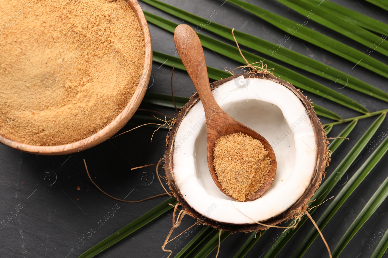 Photo of Spoon with coconut sugar, fruit, bowl and palm leaves on dark textured table, flat lay