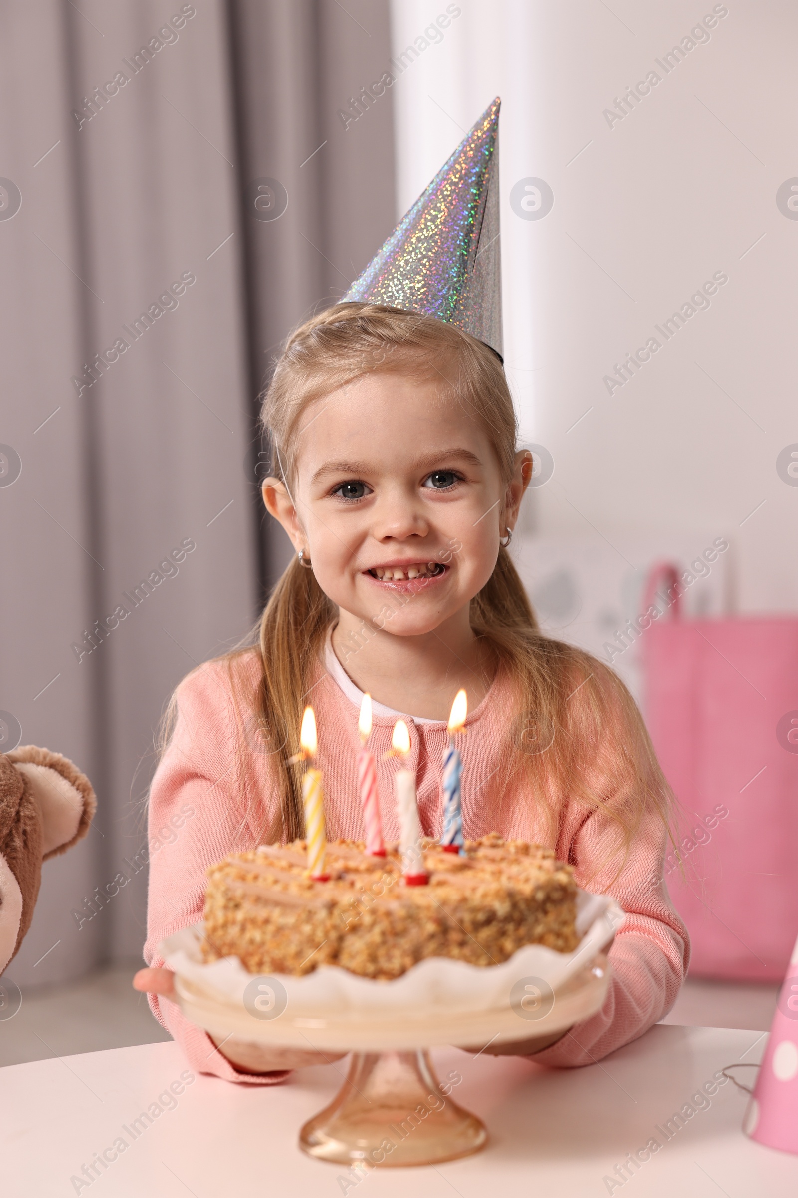 Photo of Cute girl in party hat with birthday cake at table indoors