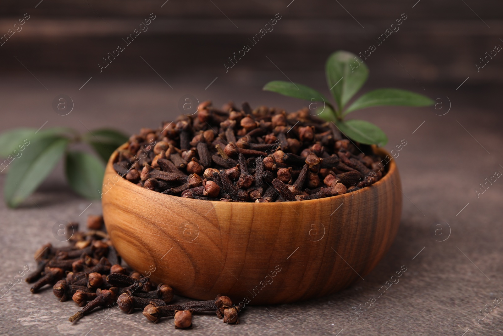 Photo of Aromatic cloves and green leaves in bowl on brown table, closeup