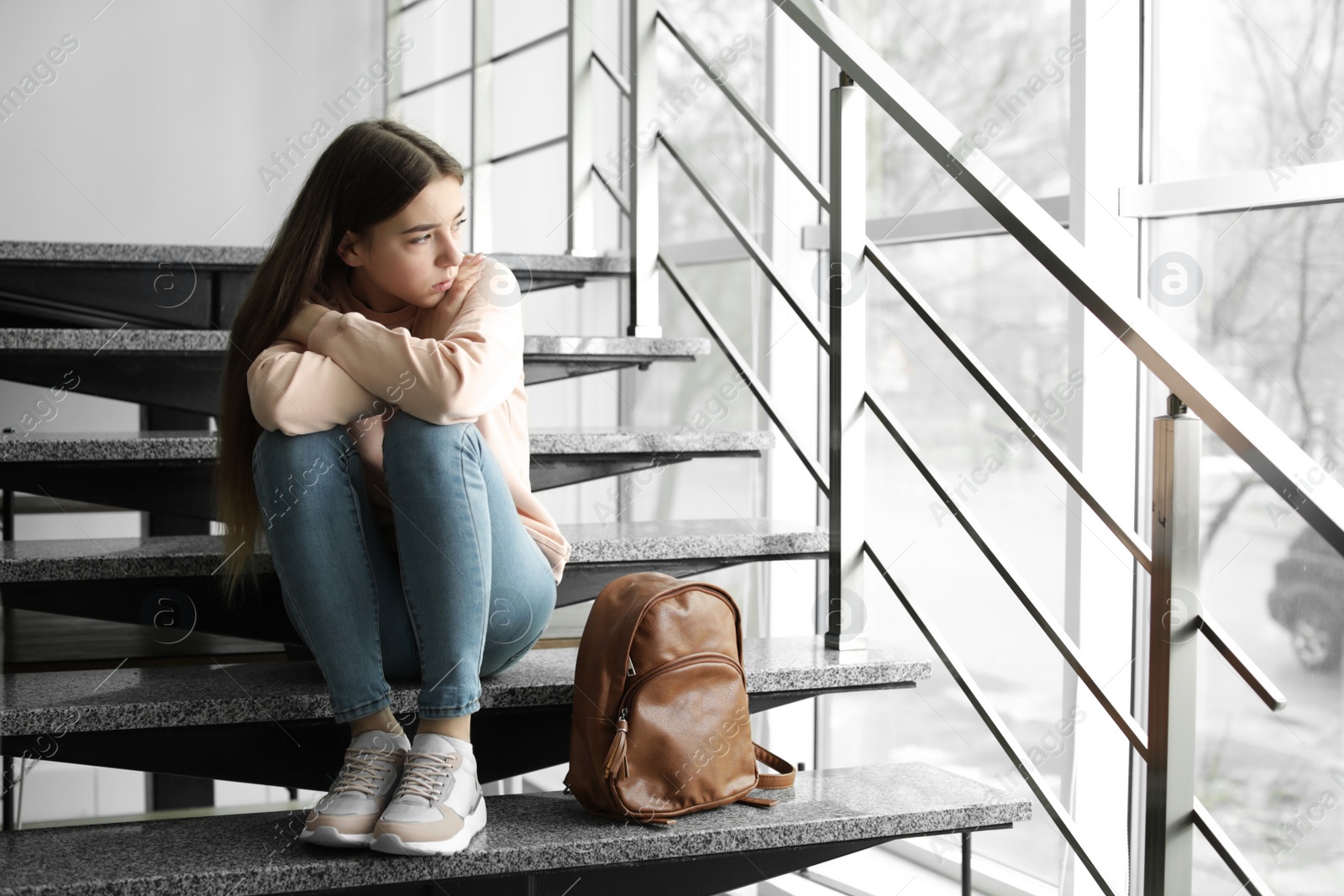 Photo of Upset teenage girl with backpack sitting on stairs indoors. Space for text