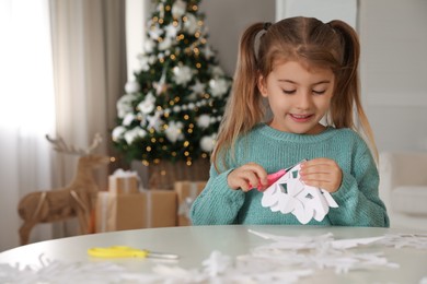 Cute little girl making paper snowflake at table near Christmas tree indoors