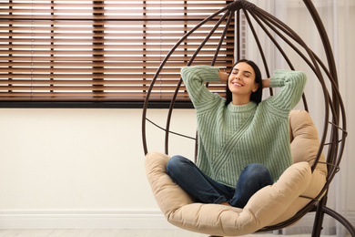 Young woman relaxing in hanging chair near window at home. Space for text