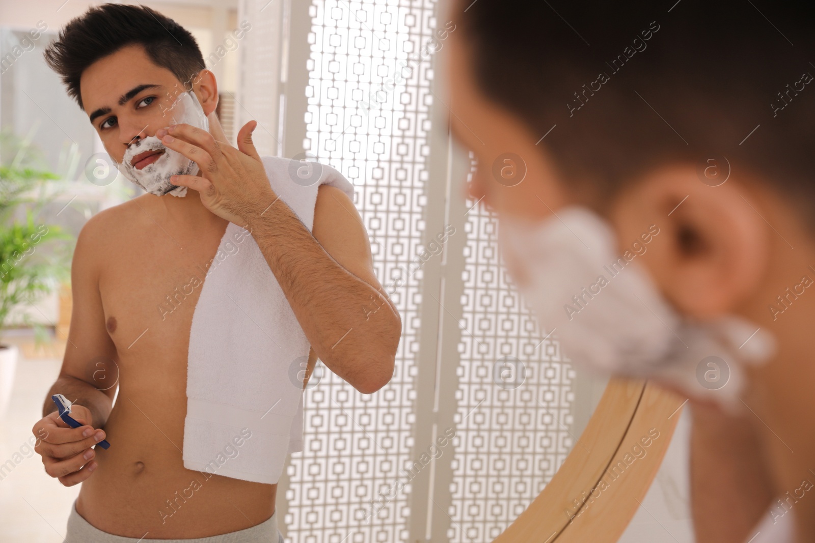 Photo of Handsome young man shaving with razor near mirror in bathroom