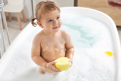 Photo of Cute little girl with sponge taking foamy bath at home