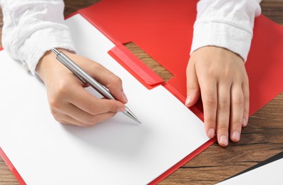 Photo of Woman writing on sheet of paper in red folder at wooden table, closeup