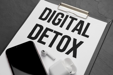 Photo of Clipboard with phrase DIGITAL DETOX, smartphone and earphones on black stone table, closeup