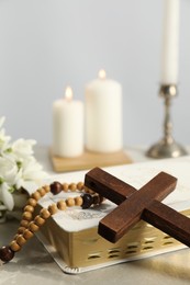 Wooden cross, Bible, rosary beads and church candles on light table, closeup