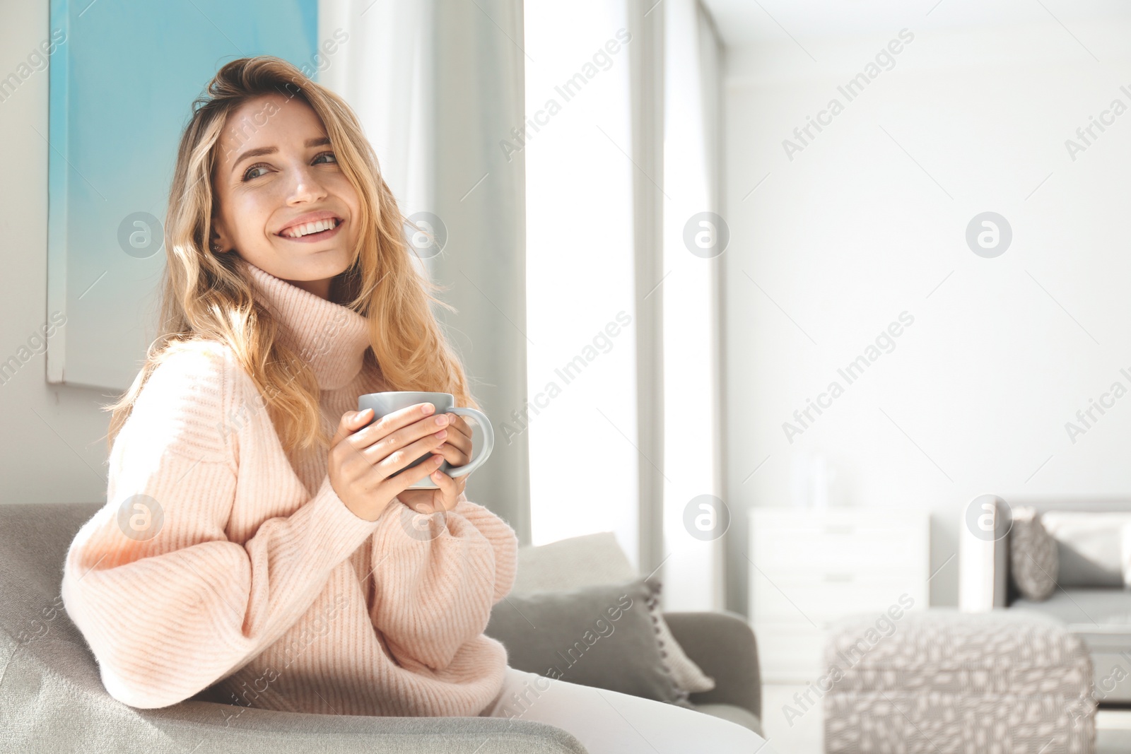 Photo of Beautiful young woman with cup of hot drink wearing warm pink sweater at home. Space for text