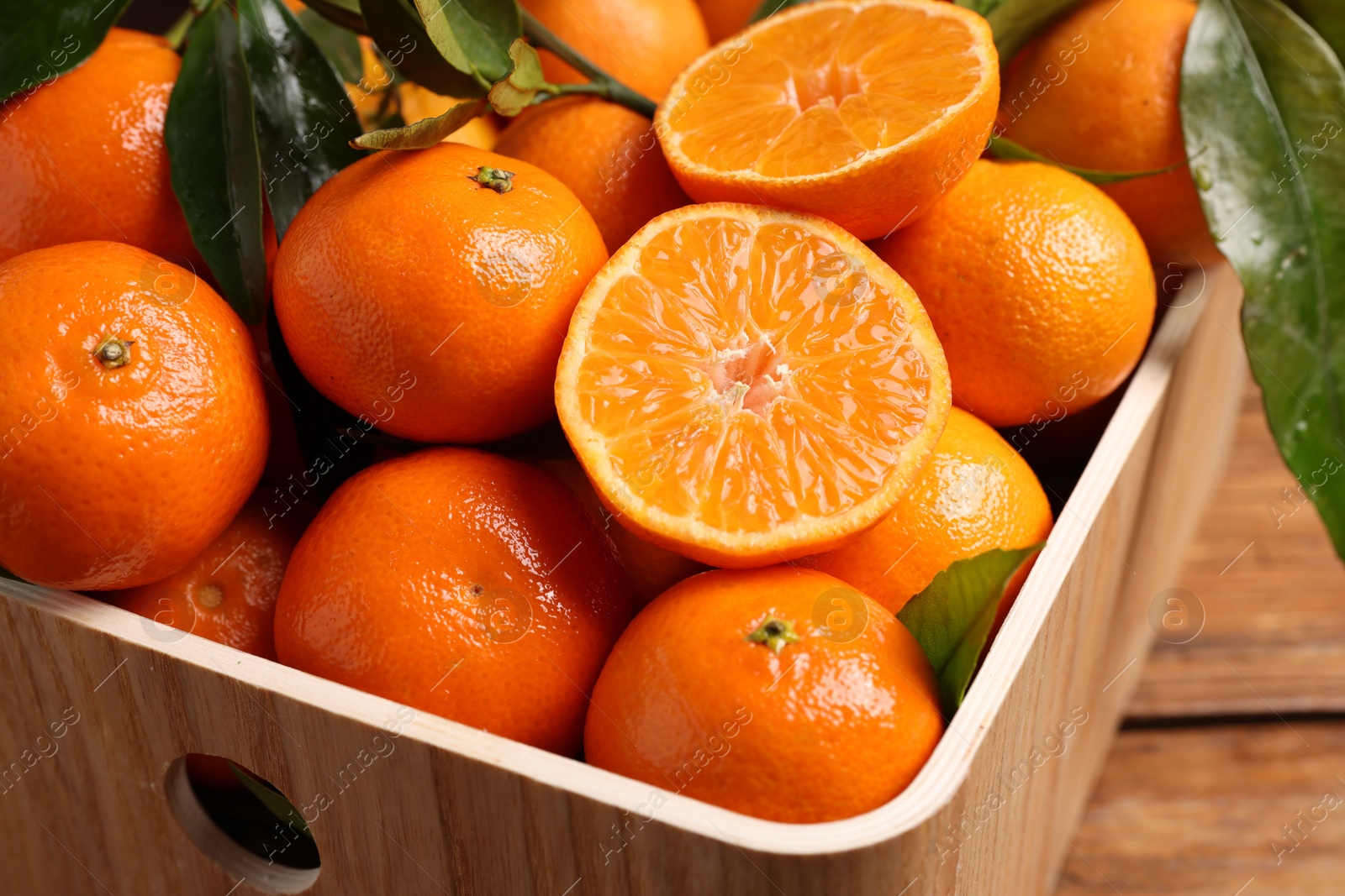 Photo of Fresh tangerines with green leaves in crate on wooden table, closeup