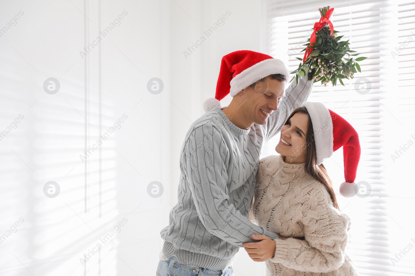 Photo of Happy couple in Santa hats under mistletoe bunch at home, space for text