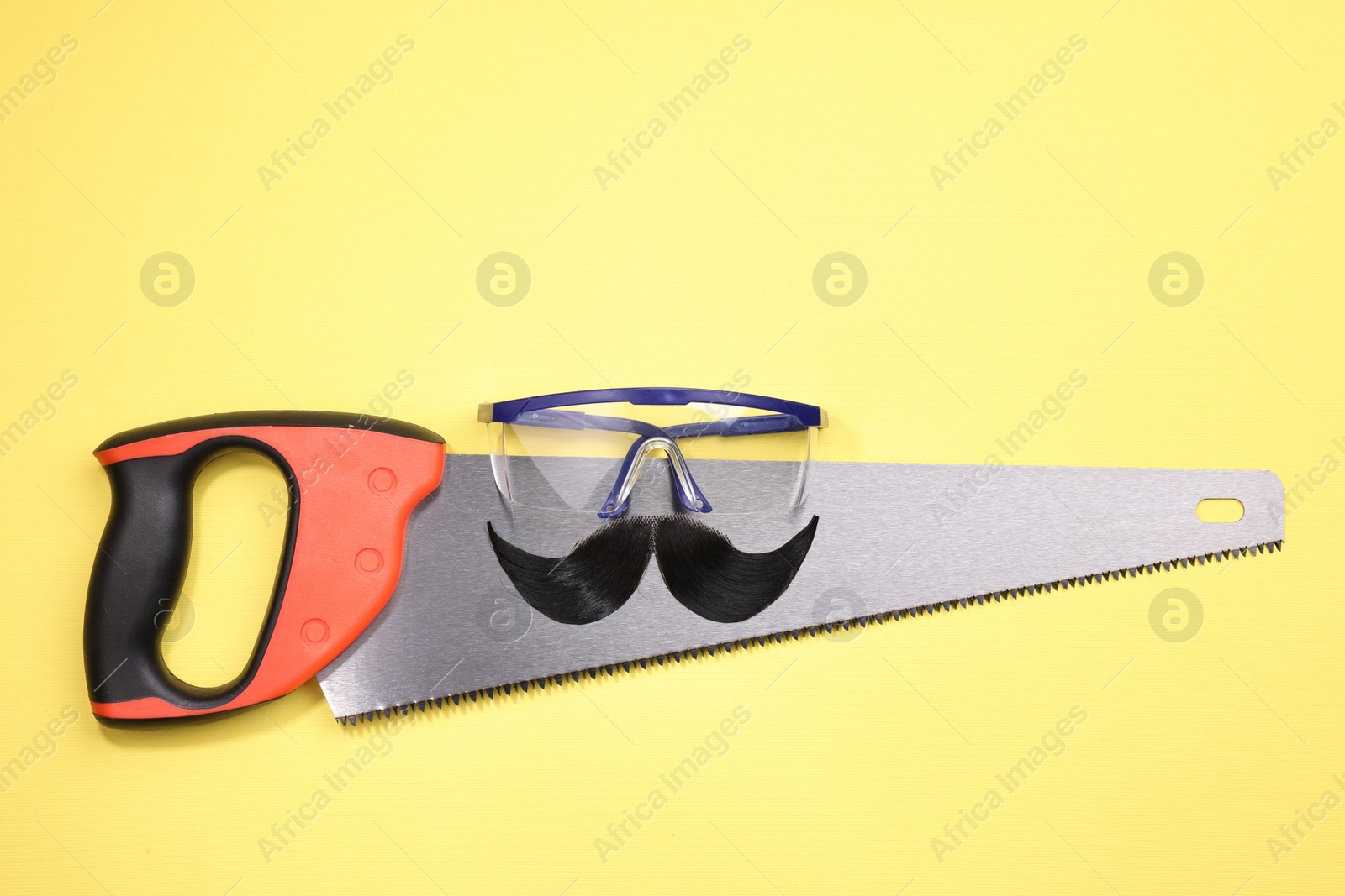 Photo of Man's face made of artificial mustache, safety glasses and hand saw on yellow background, top view