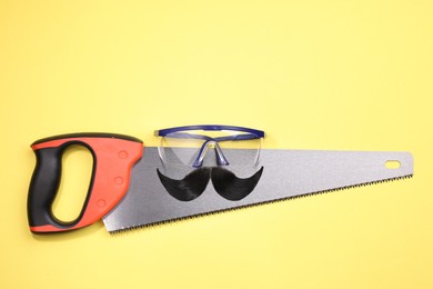 Man's face made of artificial mustache, safety glasses and hand saw on yellow background, top view