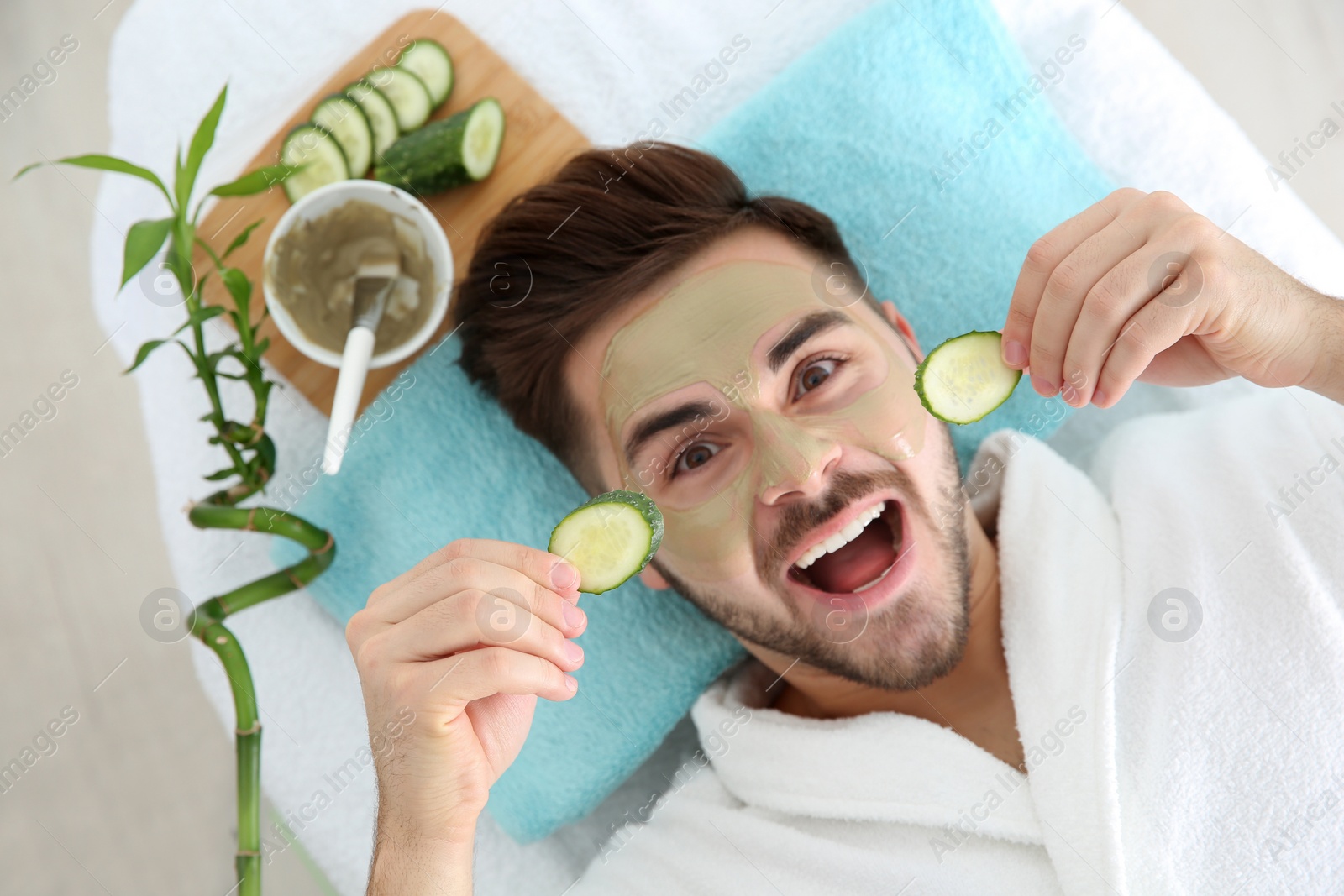 Photo of Young man with clay mask on his face holding cucumber slices in spa salon, above view