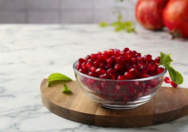 Photo of Ripe juicy pomegranate grains in bowl and green leaves on white marble table, space for text