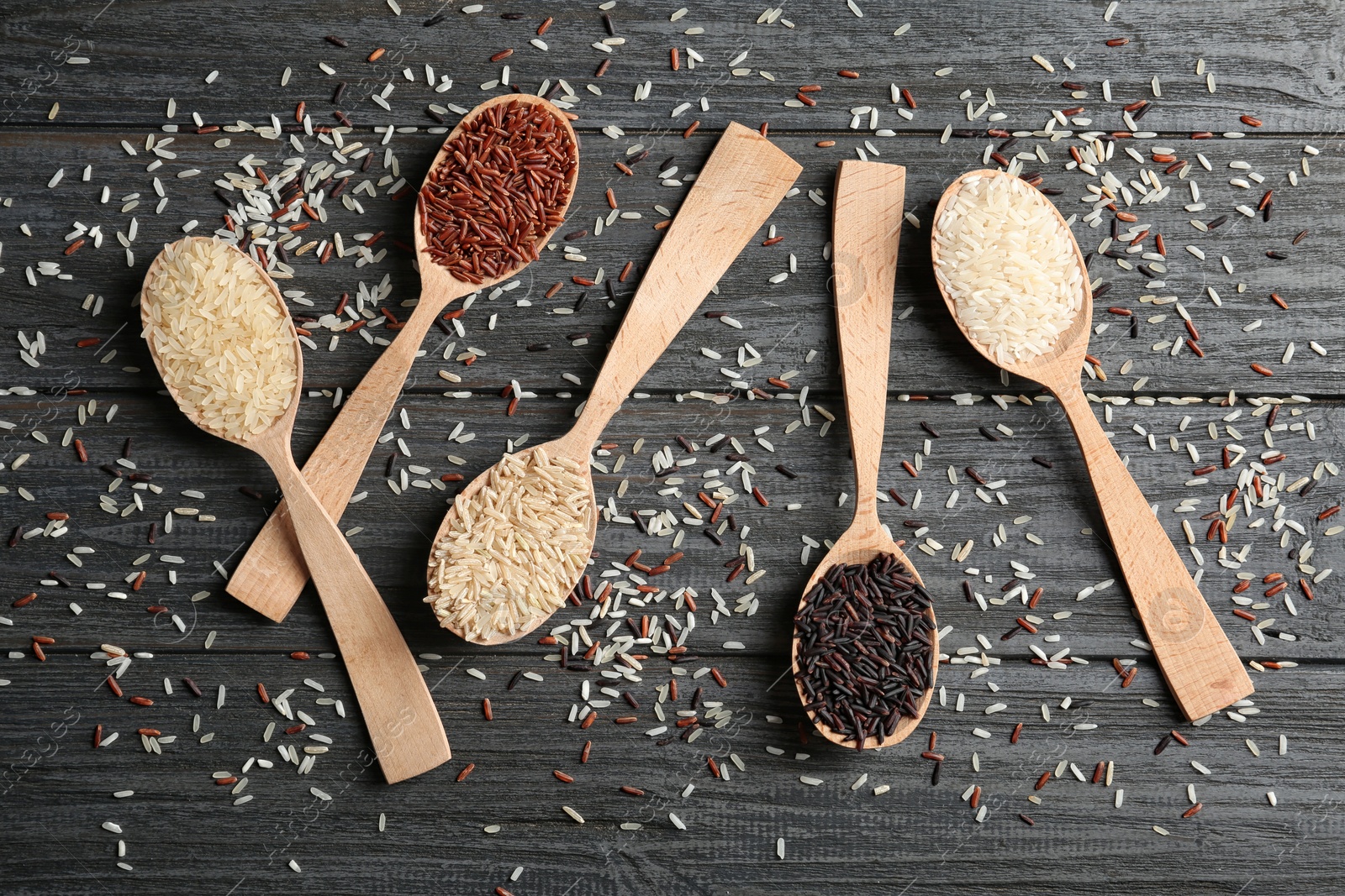 Photo of Flat lay composition with brown and other types of rice in spoons on wooden background