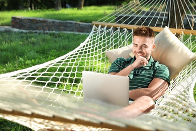 Photo of Young man with laptop resting in comfortable hammock at green garden