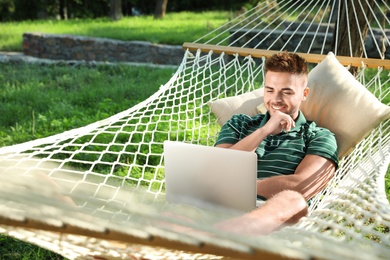 Young man with laptop resting in comfortable hammock at green garden