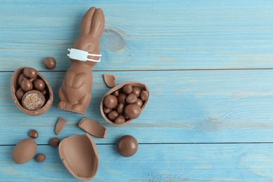 Photo of Chocolate bunny with protective mask, eggs and space for text on light blue wooden table, flat lay. Easter holiday during COVID-19 quarantine
