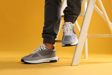 Photo of Man in stylish sneakers standing on white ladder against yellow background, closeup